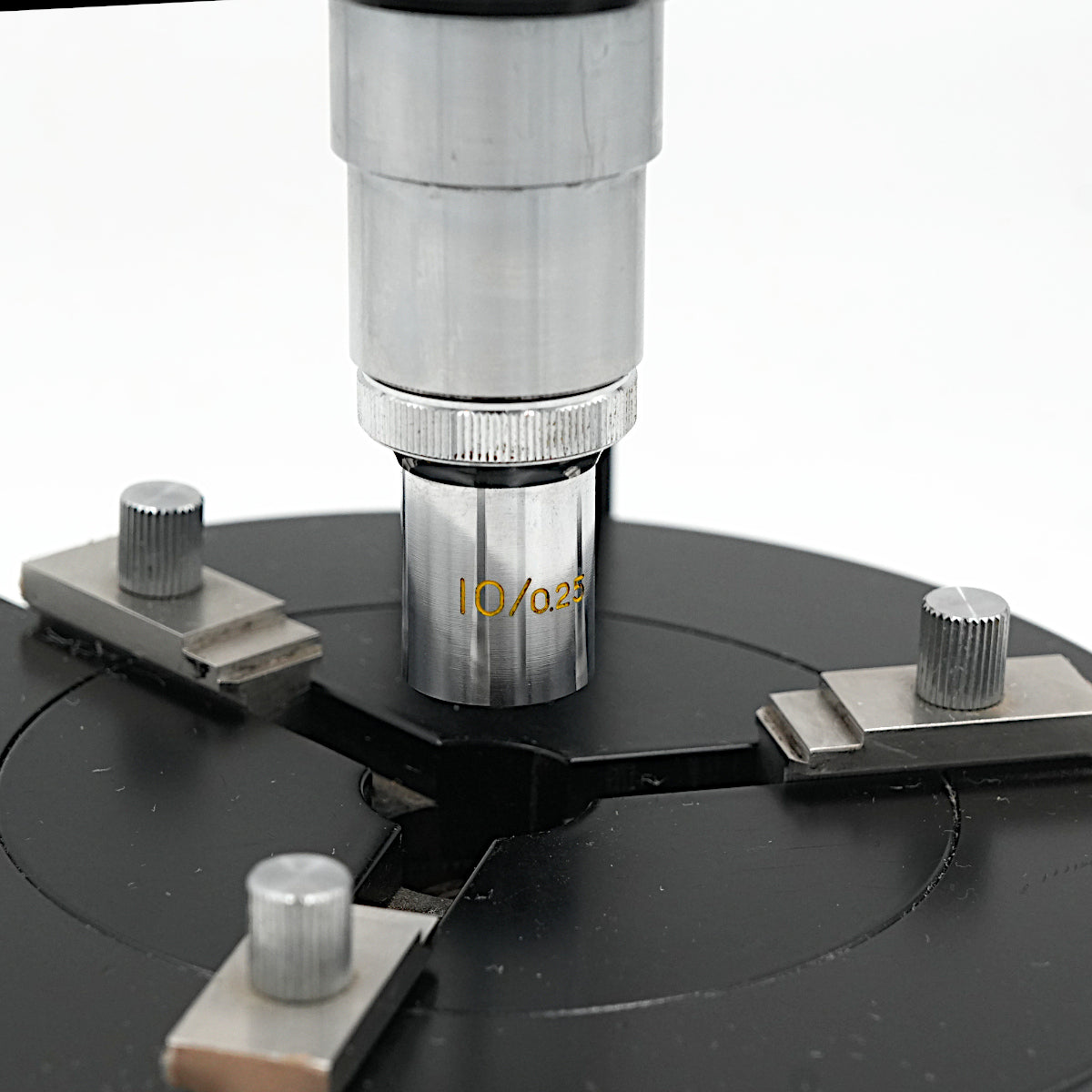Close-up of a Peal microsope objective engraved with 10/0.25 mounted on a black circular base with four adjustable silver knobs against a white background.