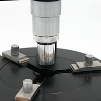 Close-up of a Peal microsope objective engraved with 10/0.25 mounted on a black circular base with four adjustable silver knobs against a white background.